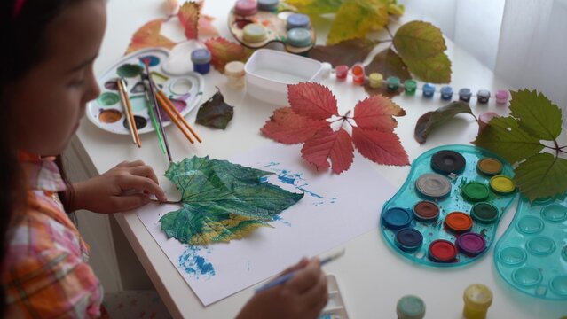 Close-up of little girl sitting at table drawing the leaves with brush and watercolours. little girl painting fallen leaves at home. autumn