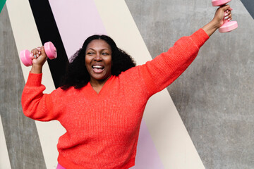 Cheerful woman with dumbbells exercising in front of wall