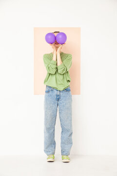 Young woman covering face with purple balloons standing against white background