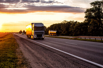 A semitrailer tractor with a tilt semitrailer transports cargo against the backdrop of an evening sunny sunset in summer. The concept of the logistics system, sanctions
