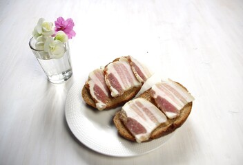 Salo or salty pork with fat slices on brown bread on plate with vadka glass and flowers on white background