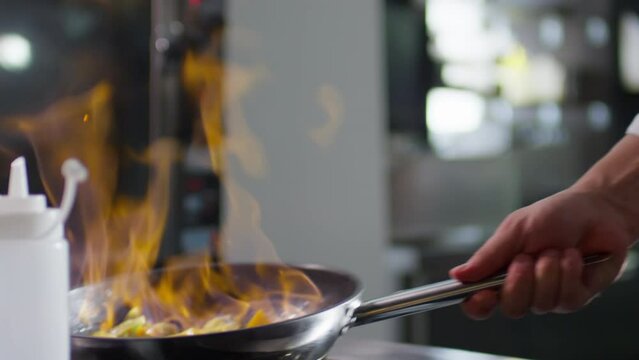 Slow motion close up shot of hands of male chef using lighter to start fire, then flambeing and tossing vegetables in skillet while cooking restaurant food