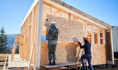 Carpenters mounting wooden OSB board on the wall of future cottage. Men workers building wooden...