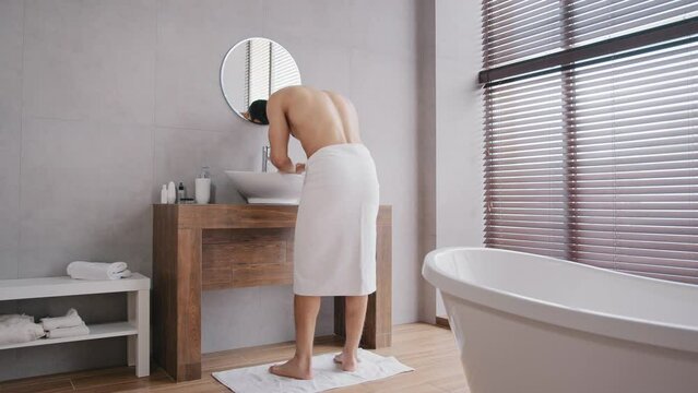 Naked bare sexy muscular Arab Indian man enters going walking in bath after shower with white towel on hips washes face with warm water in bathroom sink looks at reflection in mirror morning hygiene
