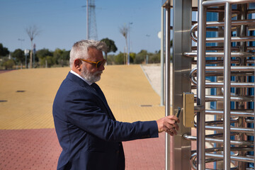 Mature, executive, gray-haired man with beard, sunglasses and jacket, showing a code with his cell phone to a reader at the entrance of the company. Concept qr codes, readers, fingerprints, security.