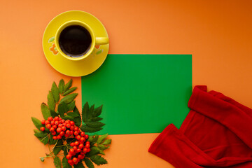 A yellow mug with a saucer, red sweater, and rowan berries on an orange background lay flat. Green sheet of paper copy space