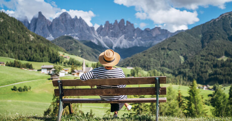 Young fashionable dressed female in straw hat sitting on a bench enjoying Santa Maddalena village view and stunning picturesque Dolomite Alps peaks in Val di Funes valley, South Tyrol, Italy. - obrazy, fototapety, plakaty