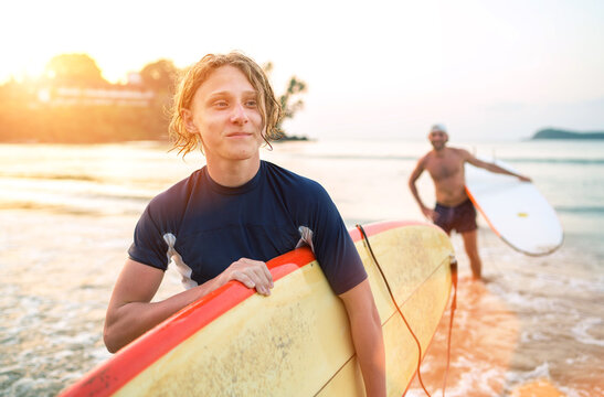 Portrait of a teen boy with a surfboard after surfing with his father. They are smiling and walking out of the water. Family active vacation concept.