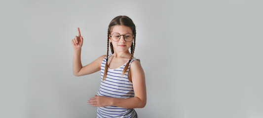 Girl child schoolgirl with pigtails in round glasses shows a finger, copy space and a light beige background