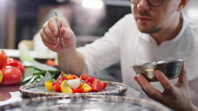 Professional chef in uniform and glasses using tweezers for garnishing fresh tomato salad with pickled red onion in restaurant kitchen