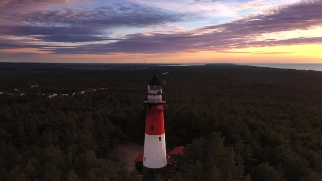 Aerial landscape of the sunset over the lush green forest. Sea in the background. Orange sky and pastel pink clouds. Flying low over the lighthouse.