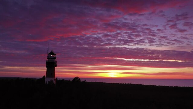 Beautiful sunset over green forest. Slow drone flight. View of the lighthouse, zoom in. Sea in the distance. Orange sun, pastel pink clouds, red sky.