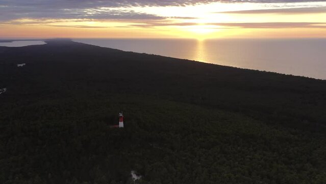 Aerial landscape of the sunset over the green forest. Sea in the background, lighthouse in the middle. Orange sky. Panoramic view. 
