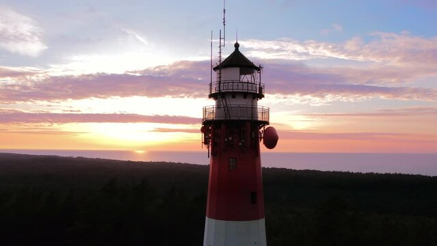 Aerial view of the lighthouse during sunset. Sea in the background. Blue sky and orange clouds. Slow drone flight over the green forest.