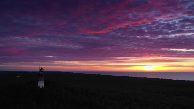 Beautiful sunset over green forest. View of the lighthouse, sea in the background. Orange sun, pastel pink clouds, red sky.