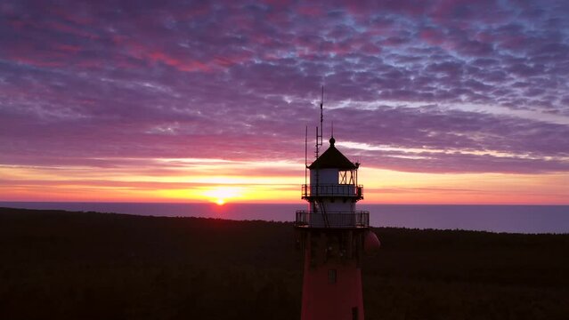 Beautiful sunset over green forest. View of the lighthouse. Orange sun, pastel pink clouds, red sky.