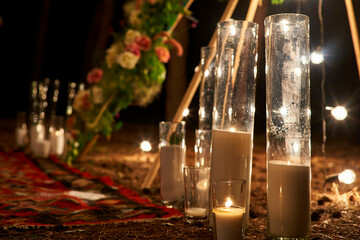 Candles burning in glass vases, flasks on carpet near bohemian tipi arch decorated with roses and flowers wrapped in fairy lights on outdoor wedding ceremony venue in pine forest. Boho rustic decor. © artiemedvedev