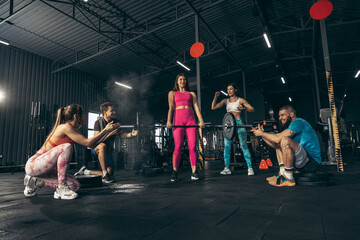 Group of young friends doing sports, training at gym indoors. Girl in bright sportswear lifting...
