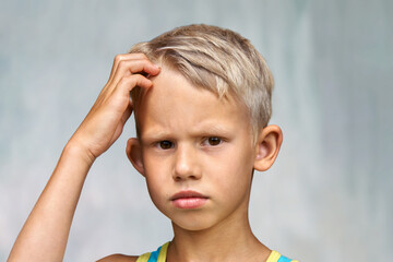 Confused junior schooler puts hand on head looking in camera. Portrait of cute blond boy with brown...