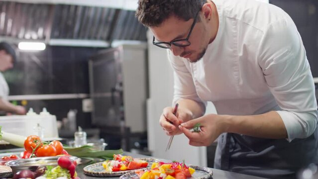 Restaurant chef in uniform and glasses adding fresh dill and pickled onion to tomato salad on plate while garnishing dish before serving