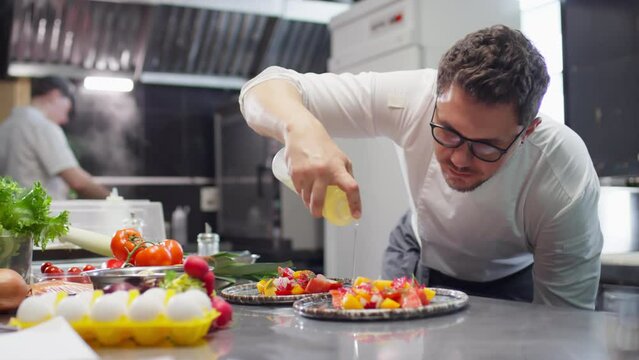 Head chef in uniform and glasses dressing tomato salad with olive oil while serving it on plate in restaurant kitchen