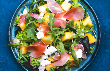 Delicious summer fresh salad with cantaloupe melon, prosciutto, soft cheese and arugula on blue table background, top view, copy space