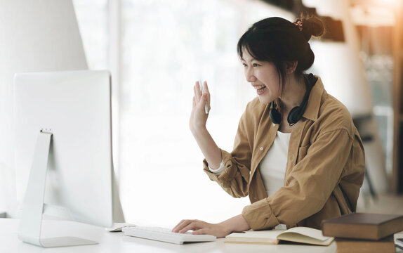 Head shot portrait smiling asian woman wearing headphones posing for photo at workplace, happy excited female wearing headset looking at laptop, sitting at desk with laptop, making video call