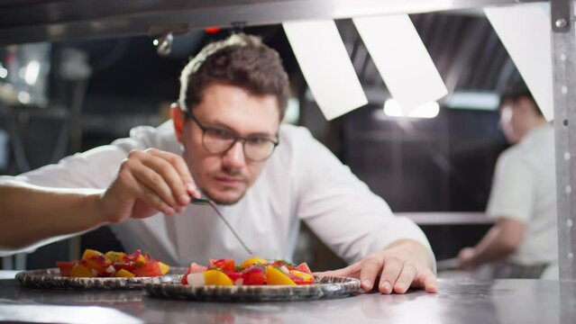 Selective focus shot of Caucasian chef in glasses and uniform serving salad on plate with tweezers at work in restaurant kitchen