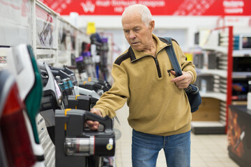 elderly grayhaired man pensioner looking Upright Vacuum hoover at counter in showroom of electrical appliance hypermarket department