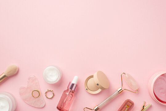Top view photo of makeup brush rose quartz roller gua sha pink eye patches glass bottles eyeshadow hairpins and gold rings on isolated pastel pink background with copyspace