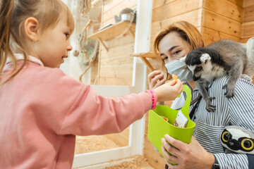 Portrait of young caucasian woman and her little daughter smiling while feeding lemur in zoo. Kid...