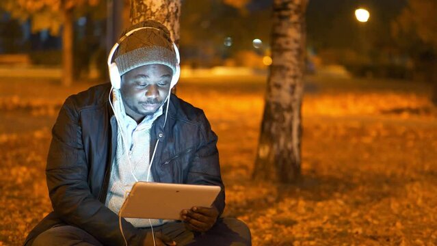 African american man in winter hat and headphones sits in evening city park and speaks via tablet computer by video link, video with flickering from lamps of artificial lighting