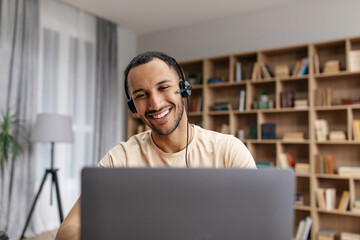 Cheerful middle eastern guy having video chat via laptop wearing wireless headphones, looking at screen and smiling