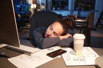 Tired young mixed race woman leaning on crossed arms while sleeping at workplace in dark office
