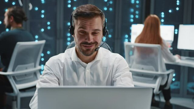 Young friendly Caucasian man with beard in headset using microphone and solving client's problem. Footage of handsome male service operator sitting at silver laptop, talking to customer via internet