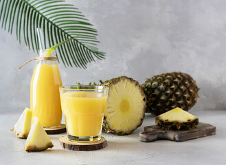 Pineapple juice in a clear glass and bottle on a light table. Half a pineapple and slices in the...