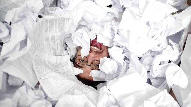 Face of male office worker screaming buried under pile of crumpled papers, top view. Man screams from under pile crumpled papers