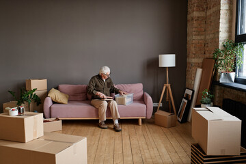 Horizontal long shot of modern senior Caucasian man sitting on sofa in living room unpacking moving to new house