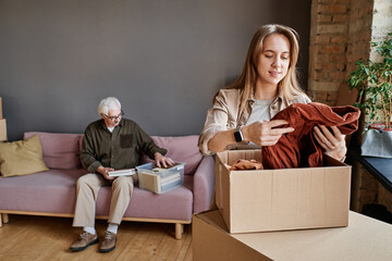 Portrait of young woman helping her grandfather to unpack clothes and stuff after moving to another apartment - obrazy, fototapety, plakaty