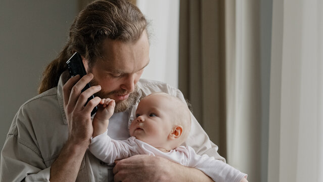 Cheerful Carefree Middle-aged Caucasian Man Bearded Father Answering Call Talking To Wife Mom With Phone Online Order Booking Baby Food Holding Little Daughter Newborn Infant At Home Chatting Friendly