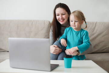 Fototapeta premium Mother and her daughter using video chat on laptop sitting on cozy sofa at home, watching video, having fun, two generations