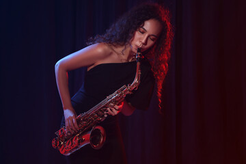 Beautiful African American woman playing saxophone on dark background - obrazy, fototapety, plakaty