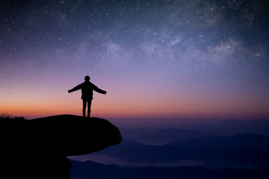 Silhouette of young traveler standing and open arms alone on top of the mountain and watched the beautiful view night sky, star and milky way. He was happy to travel alone in the wide world.