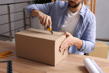 Man using utility knife to open parcel at wooden table indoors, closeup