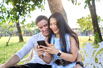Smiling couple using the mobile while sitting on a bench outdoors during sunset
