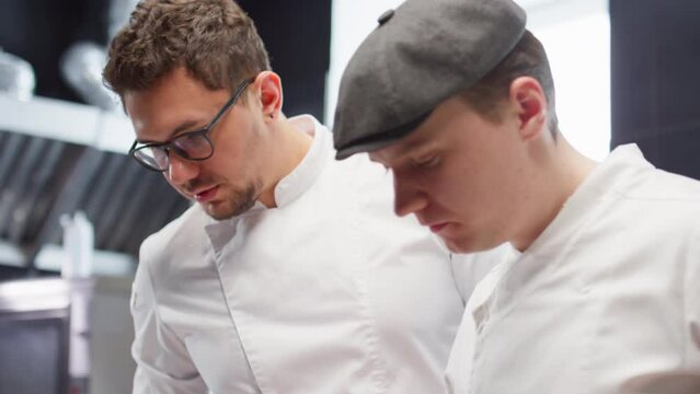 Sous chef serving cooked fish with veggies on side on plates as head chef teaching him while working together in restaurant kitchen