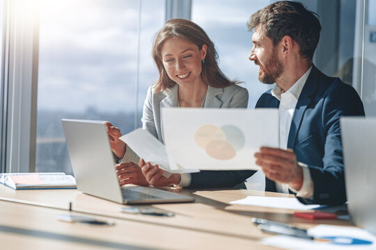 Two Business People During Work Meeting In Modern Office Explaining Statistics Data To Each Other Looking At Documents With Graphs And Charts