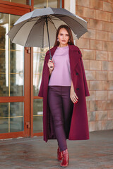 A model girl showing fashionable outerwear with an umbrella in her hand, on the street, on the veranda of a summer cafe. Clothes for the showroom.