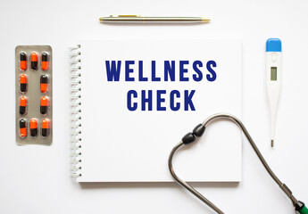 WELLNESS CHECK is written in a notebook on a white table next to pills and a stethoscope.