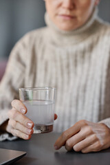Vertical shot of mature woman having cold wearing sweater sitting at desk at home holding glass with mixture of water and effervescent tablet